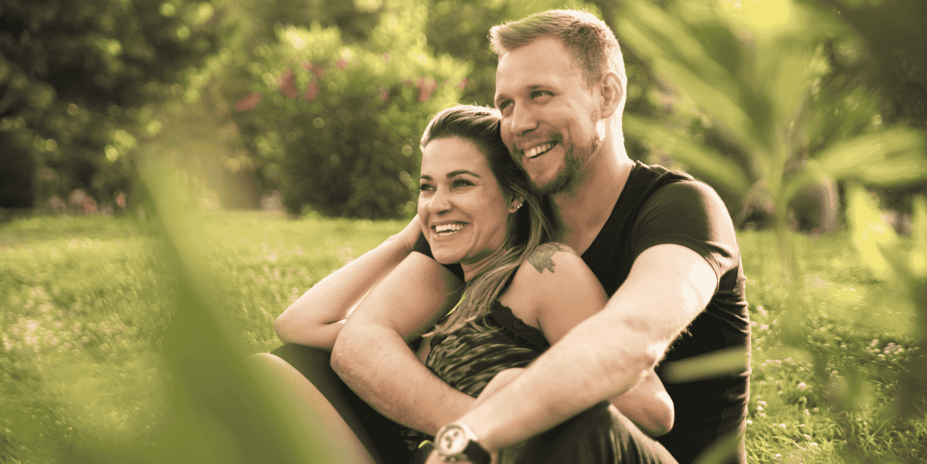 Couple on a date in a park sitting on the ground and hugging with the woman sitting in the man's lap and both are smiling.